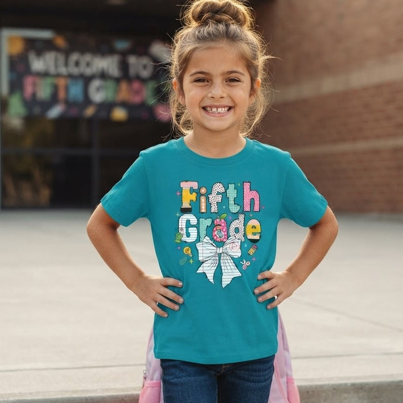 A young girl smiling on outdoor concrete steps in front of a brick building, wearing a teal short-sleeve t-shirt with a colorful 'Fifth Grade' graphic and a white bow design on the front, paired with dark blue jeans.