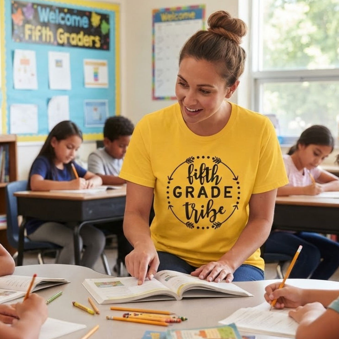 A woman wearing a bright yellow graphic t-shirt with the text 'fifth grade tribe' sits at a desk in a classroom, smiling and pointing to an open book. Other students are visible in the background.