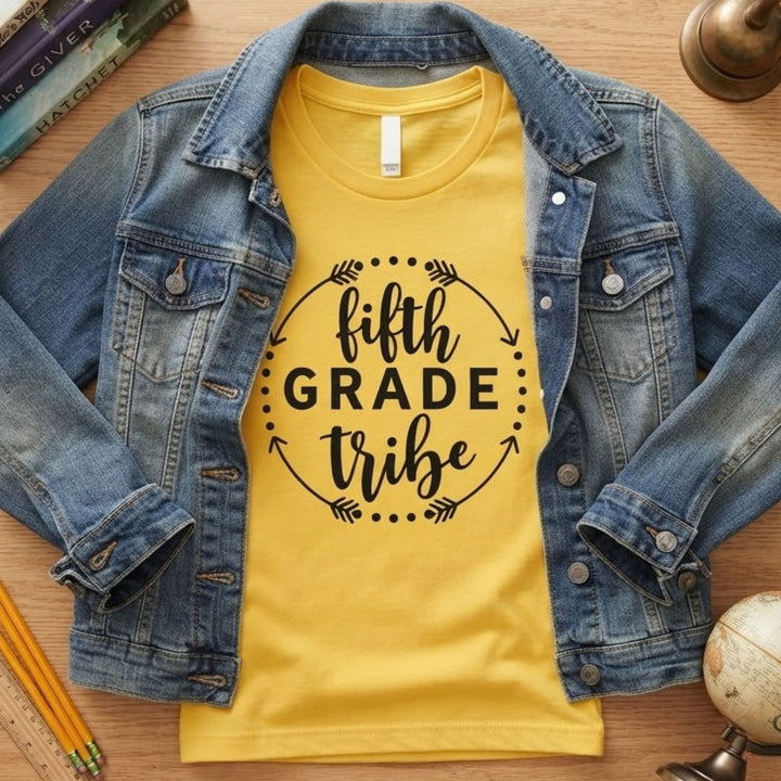 A yellow graphic t-shirt with a black 'fifth grade tribe' print, layered under an open blue denim jacket, presented in a flat-lay style on a wooden desk with school supplies.