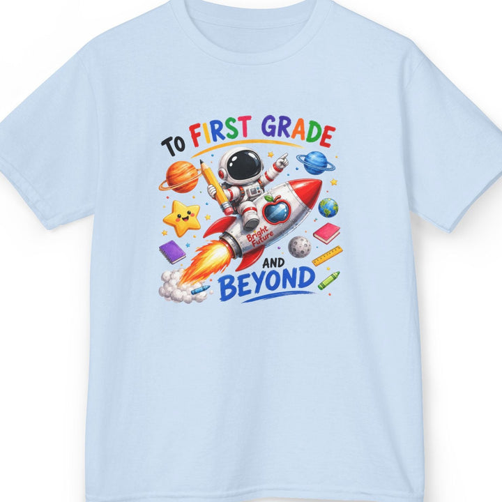 A light blue short-sleeve crewneck t-shirt featuring a colorful graphic of an astronaut riding a rocket ship with the text 'TO FIRST GRADE AND BEYOND' on a white background.