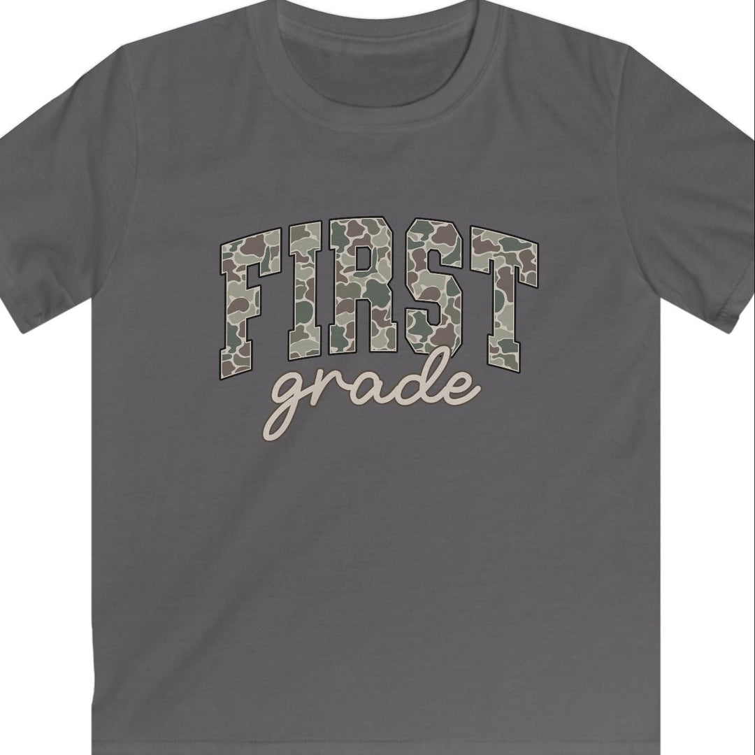 A dark gray short-sleeve t-shirt with 'FIRST grade' printed on the chest. The word 'FIRST' is in a camouflage pattern, and 'grade' is written below in a light beige cursive font, set against a plain white background.