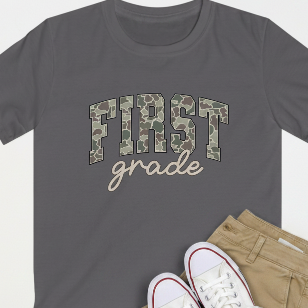 A dark charcoal gray crewneck t-shirt featuring a 'FIRST grade' graphic on the chest, styled in a flat lay with khaki pants and white sneakers on a light gray background.