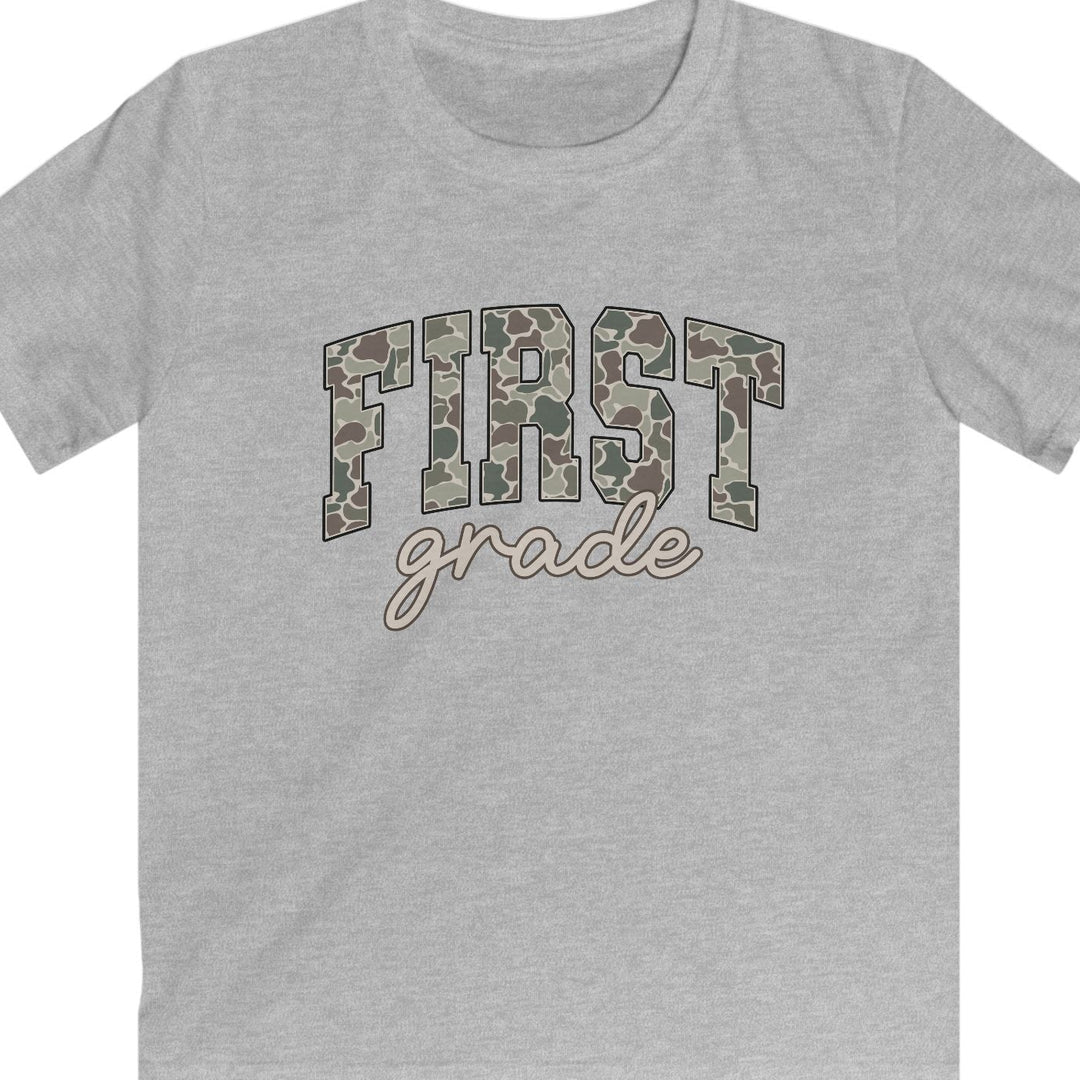 A heather gray short-sleeve t-shirt with a graphic on the chest reading 'FIRST grade' in camouflage block letters and cursive script, shown on a plain white background.