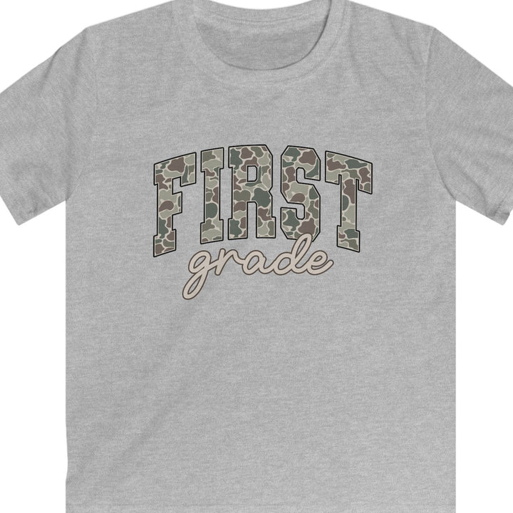 A heather gray short-sleeve t-shirt with a graphic on the chest reading 'FIRST grade' in camouflage block letters and cursive script, shown on a plain white background.