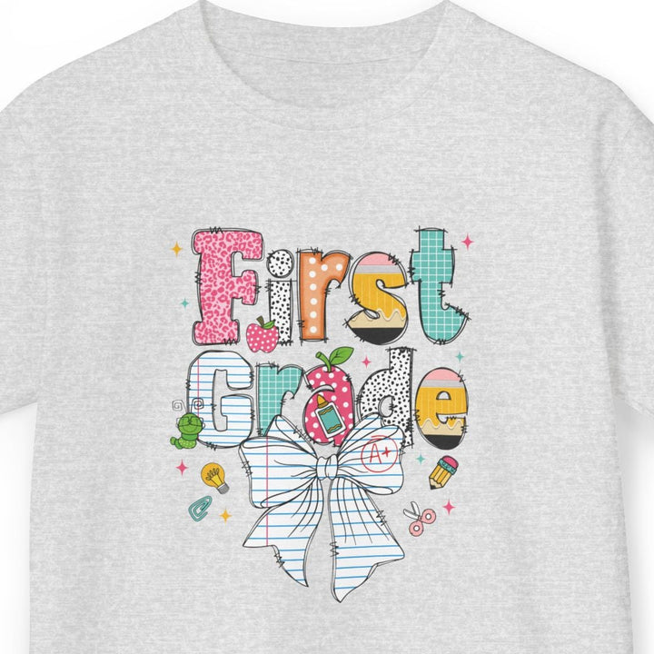 A light ash gray short-sleeve t-shirt with a colorful graphic on the front that reads 'First Grade' decorated with a striped bow, candy, and school-themed doodles, shown against a plain white background.