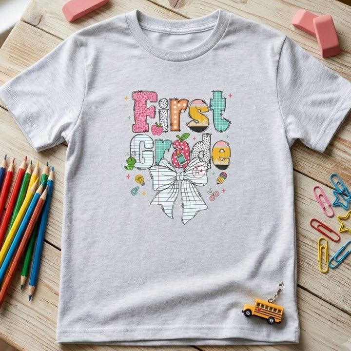 A light gray heathered short-sleeve t-shirt with a colorful 'First Grade' school-themed graphic, styled in a flat-lay on a wooden table with school supplies.