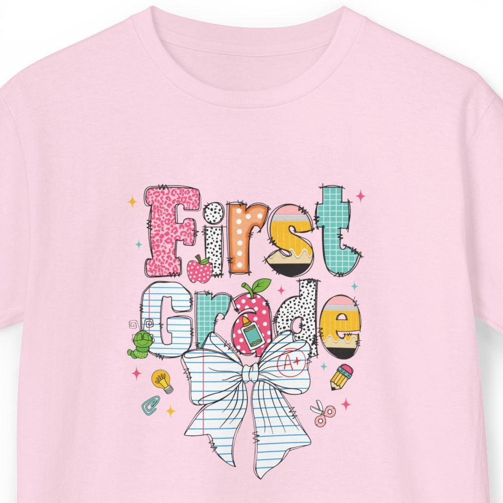 A light pink short-sleeve t-shirt with a colorful graphic on the front that reads 'First Grade' decorated with a striped bow, candy, and school icons, shown on a plain white background.