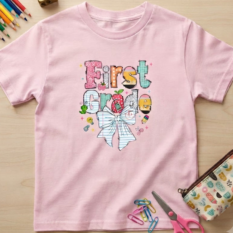 A light pink short-sleeve t-shirt with a colorful 'First Grade' graphic featuring a bow and school-themed illustrations, laid flat on a light wood background with school supplies.