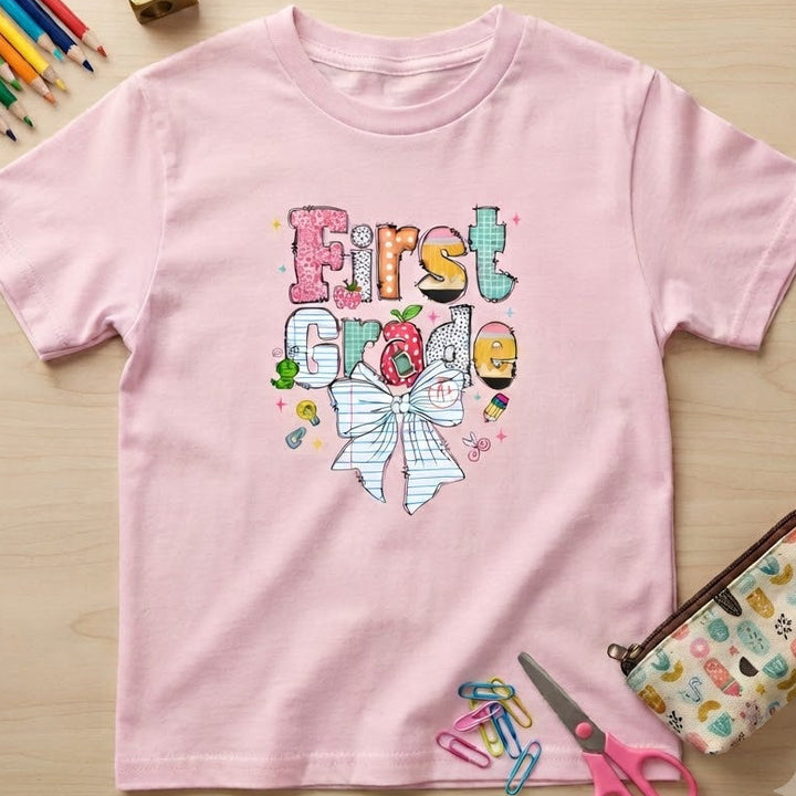 A light pink short-sleeve t-shirt with a colorful 'First Grade' graphic featuring a bow and school-themed illustrations, laid flat on a light wood background with school supplies.