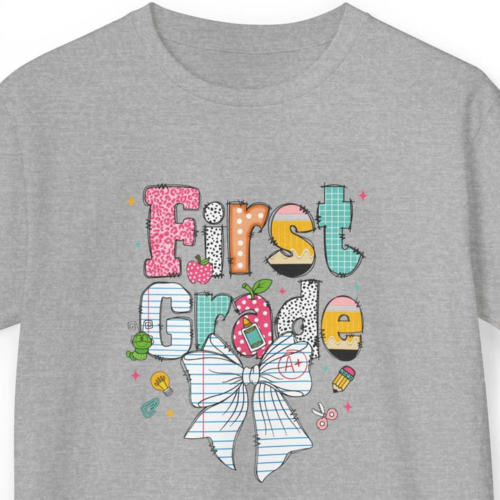 A heather gray short-sleeve t-shirt with a colorful graphic on the front that reads 'First Grade' decorated with a striped bow, a pink polka-dot hair clip, and small school-themed doodles, shown against a plain white background.