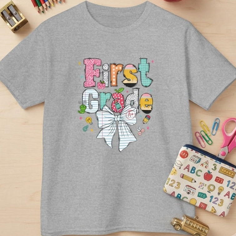 A heather gray short-sleeve t-shirt with a colorful 'First Grade' graphic featuring a striped bow and school-themed doodles, styled in a flat lay on a light wood background with school supplies and a red apple.