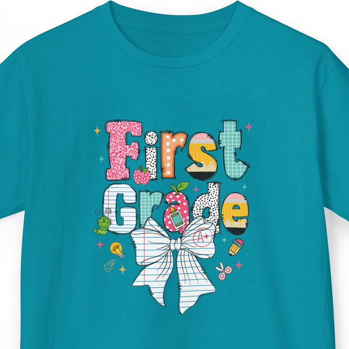 A teal short-sleeve t-shirt with a colorful 'First Grade' graphic and a white bow illustration on the front, displayed against a plain white background.