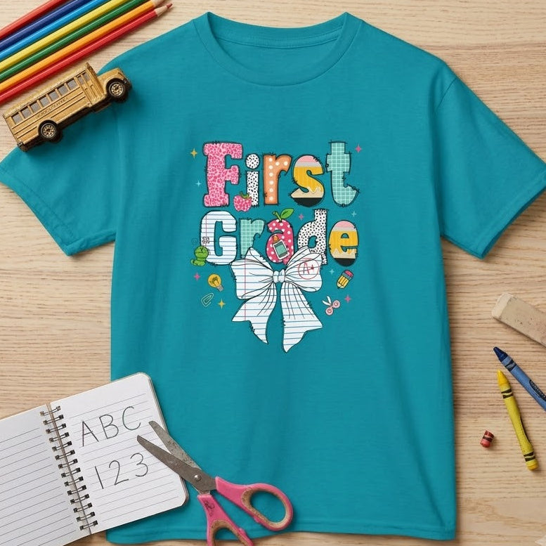 A turquoise short-sleeve T-shirt laid flat on a light wood surface, printed with colorful 'First Grade' text and a white bow graphic. Surrounding props include colored pencils, a toy school bus, a notebook with 'ABC 123', scissors, and crayons.