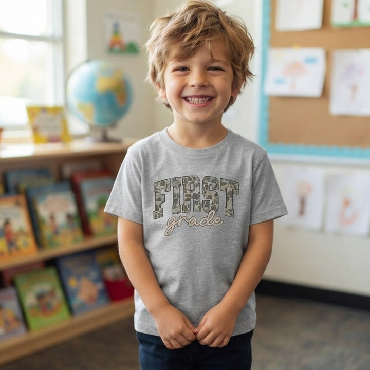 A young boy smiling in a classroom, wearing a light gray heather t-shirt with 'FIRST grade' printed on the front in camouflage and tan lettering, paired with dark blue jeans and red sneakers.