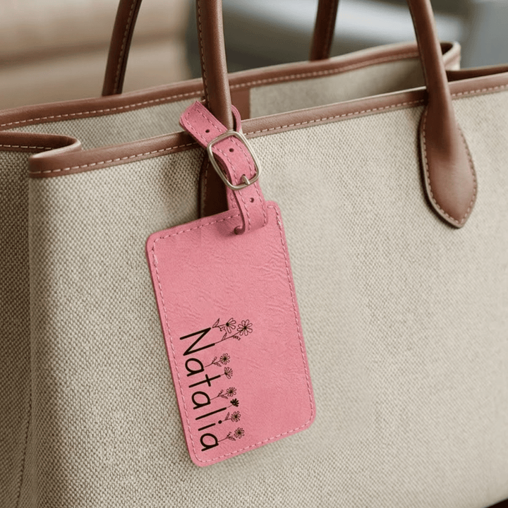 Beige handbag with a pink luggage tag featuring the name 'Natalia' engraved in black with floral accents on tops of letters.