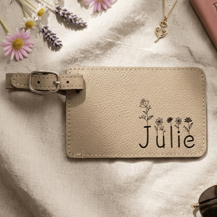 Beige leather luggage tag with 'Julie' and floral design engraved in black laying on a textured surface with flowers and jewelry.