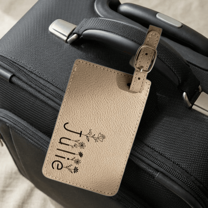 Beige luggage tag with 'Julie' and floral designs engraved in black and attached to a black suitcase.