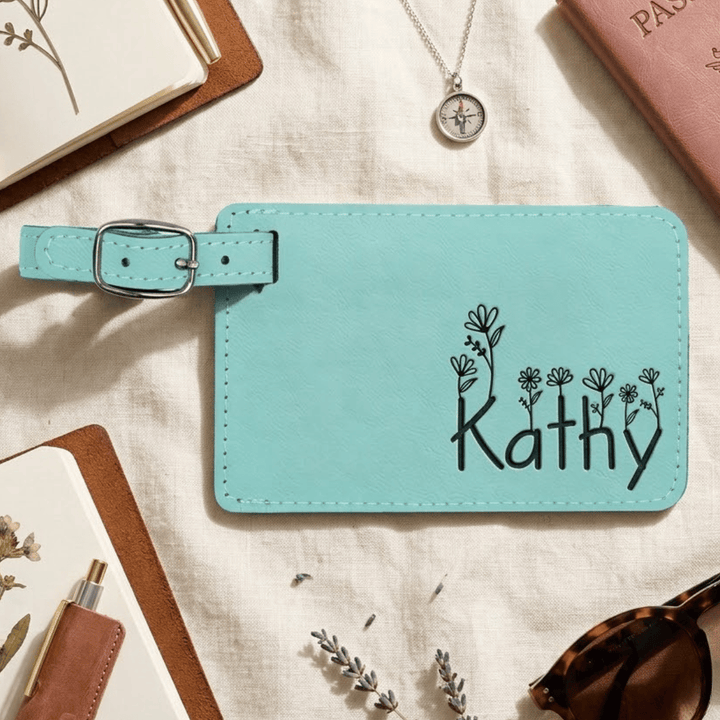Teal luggage tag with 'Kathy' and floral design engraved in black laying on a textured surface with accessories.