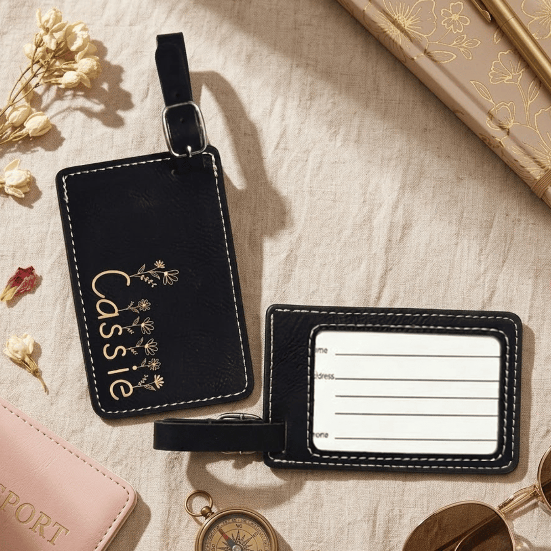 Front and back of black leather luggage tag with 'Cassie' and floral design engraved in metallic gold on a beige surface with travel items.