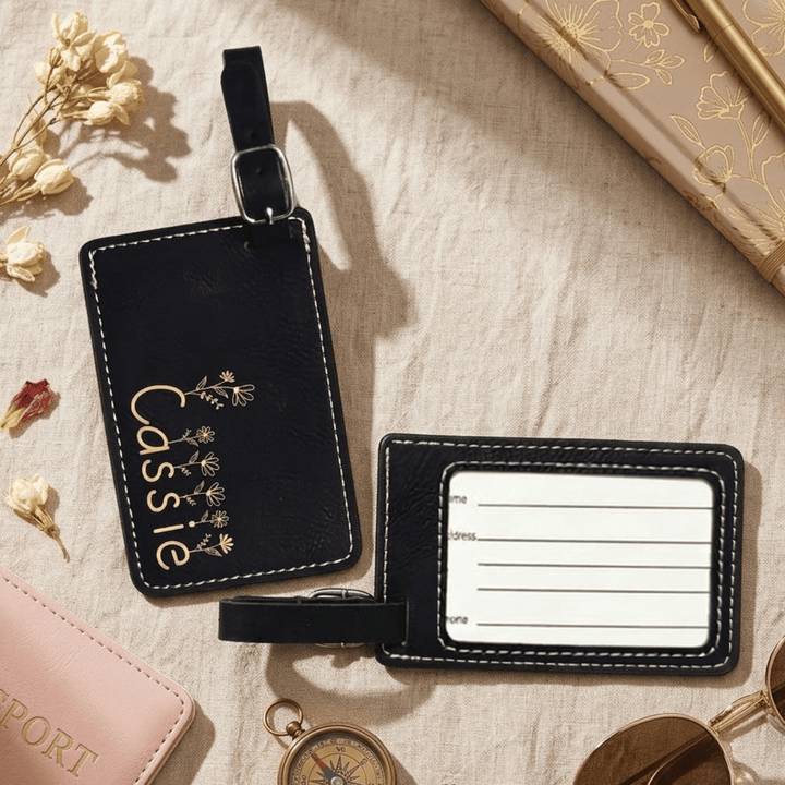 Front and back of black leather luggage tag with 'Cassie' and floral design engraved in metallic gold on a beige surface with travel items.