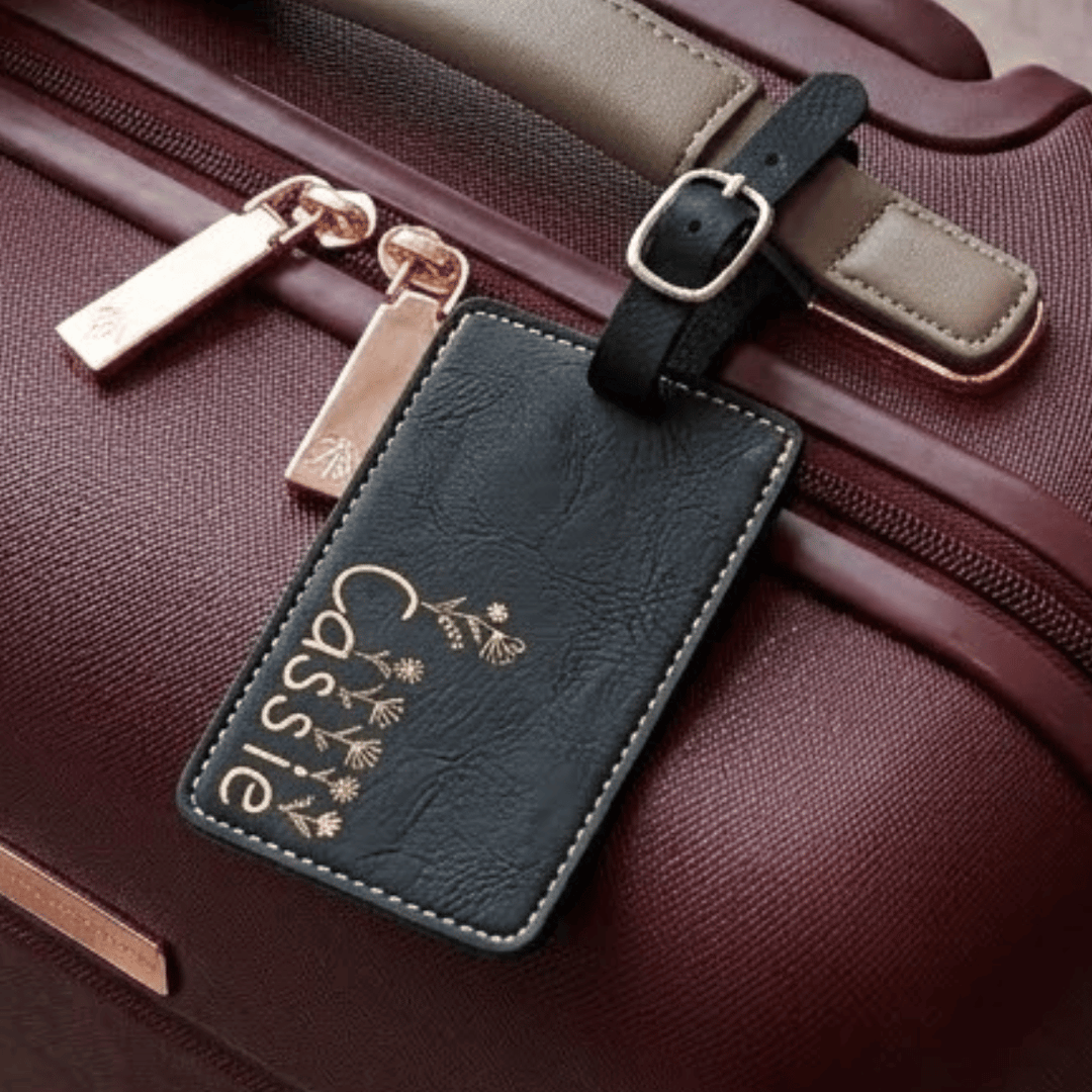 Black leather luggage tag with 'Cassie' and floral design engraved in metallic gold on a brown suitcase.