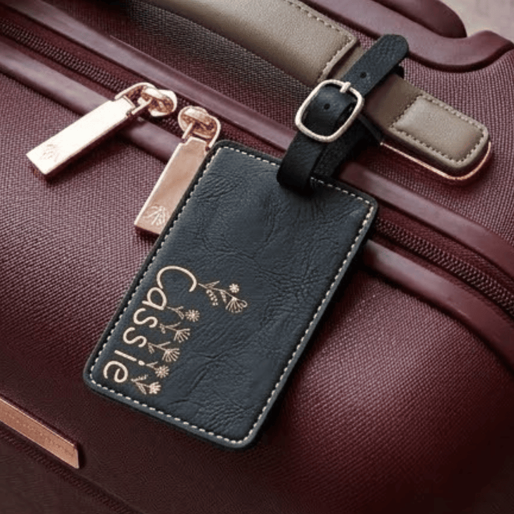 Black leather luggage tag with 'Cassie' and floral design engraved in metallic gold on a brown suitcase.