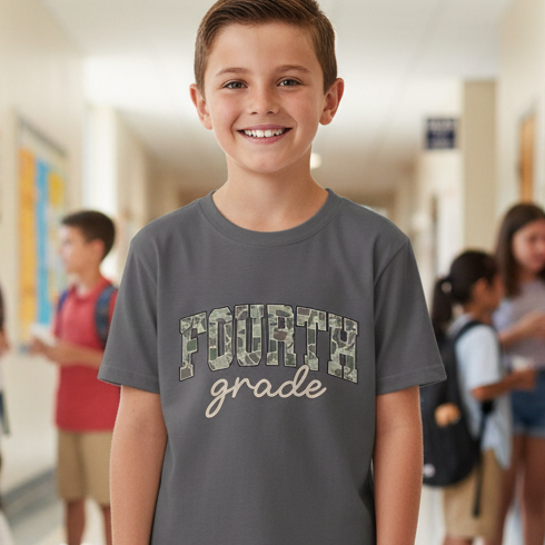 A smiling young boy stands in a school hallway wearing a dark gray t-shirt with 'FOURTH grade' printed on the front. The letters in 'FOURTH' are filled with a camouflage pattern, and 'grade' is written in a light beige script.