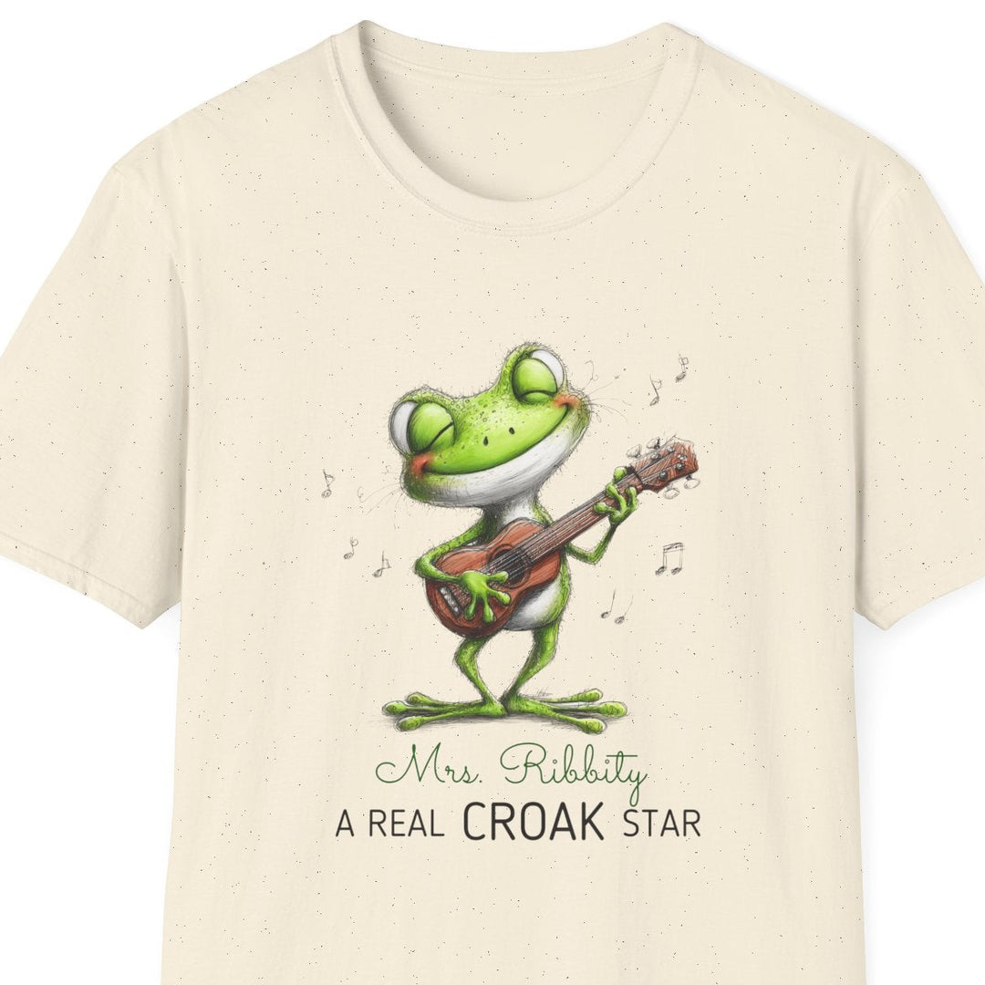 A cream-colored short-sleeve t-shirt with a cartoon frog playing a guitar on the front, accompanied by the text 'Mrs. Ribbity A REAL CROAK STAR'.