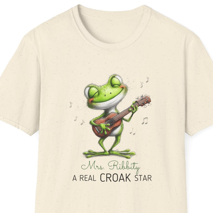 A cream-colored short-sleeve t-shirt with a cartoon frog playing a guitar on the front, accompanied by the text 'Mrs. Ribbity A REAL CROAK STAR'.