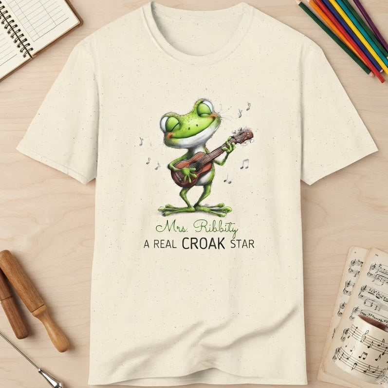 A cream-colored short-sleeve t-shirt with a cartoon frog playing a guitar and the text 'Mrs. Ribbity A REAL CROAK STAR' printed on the front.