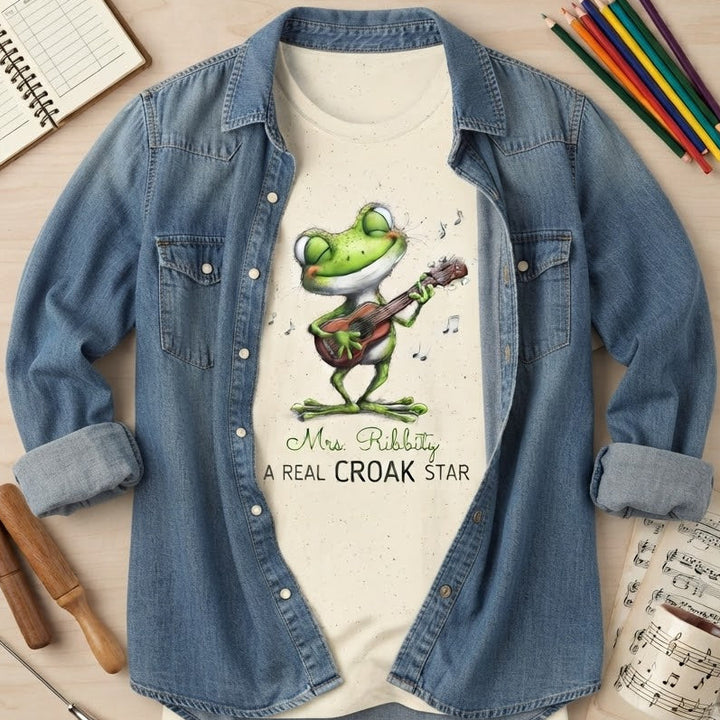 A styled flat-lay of a graphic t-shirt under an open blue denim shirt. The t-shirt features a green cartoon frog playing a guitar with the text 'Mrs. Ribbity a real croak star'.