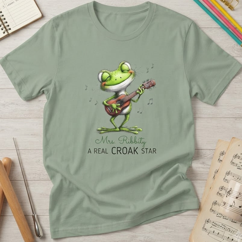 A sage green short-sleeve t-shirt with a cartoon frog playing a guitar and the text 'Mrs. Ribbity A REAL CROAK STAR' printed on the front.