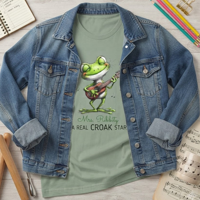 A styled flat-lay of a light sage green graphic T-shirt under an open blue denim jacket. The shirt features a cartoon frog playing a guitar with the text 'Mrs. Ribbity A REAL CROAK STAR'.