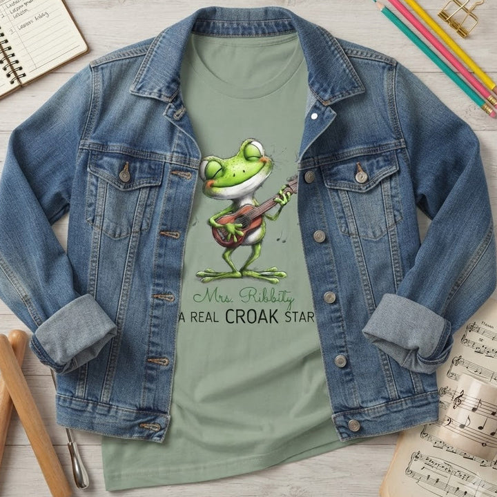 A styled flat-lay of a light sage green graphic T-shirt under an open blue denim jacket. The shirt features a cartoon frog playing a guitar with the text 'Mrs. Ribbity A REAL CROAK STAR'.