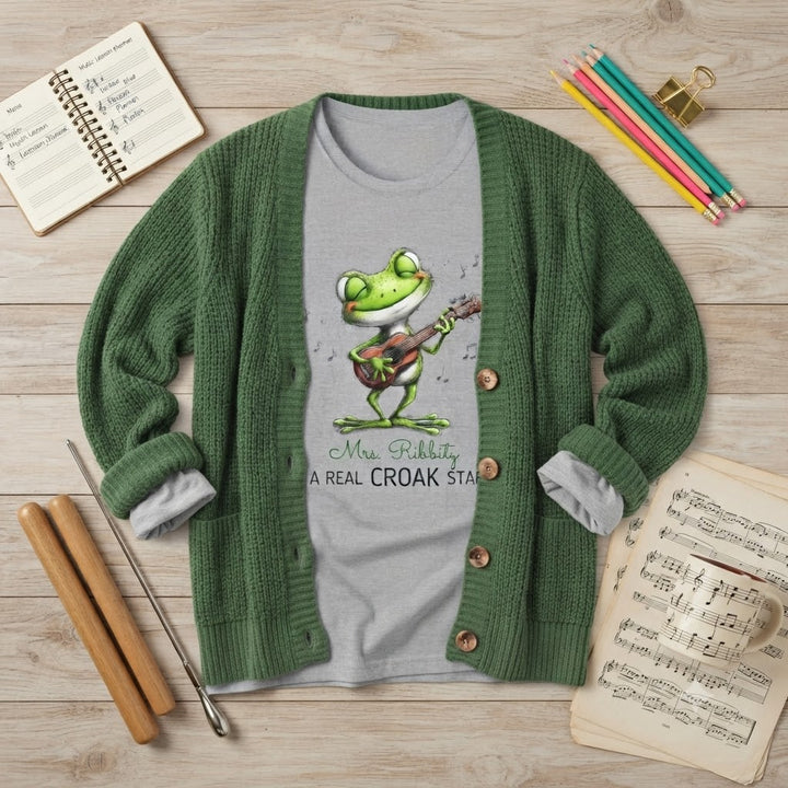 A green knit cardigan sweater laid flat on a light wood surface, worn open over a light gray T-shirt featuring a cartoon frog playing a guitar with the text 'Mr. Ribbity, A REAL CROAK STAR'.