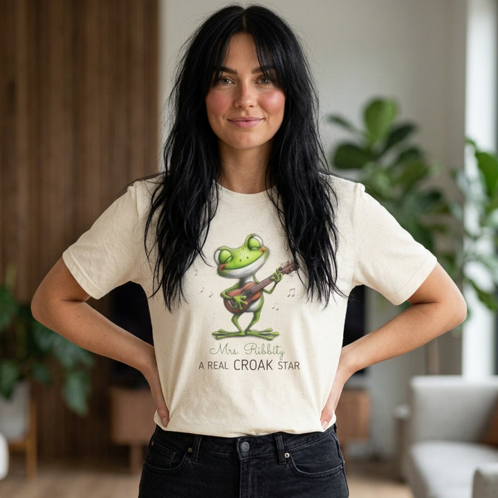 A woman with long black hair stands indoors wearing a cream-colored graphic T-shirt with a green frog playing guitar and the text 'Mrs. Rabbit A REAL CROAK STAR', paired with black jeans.