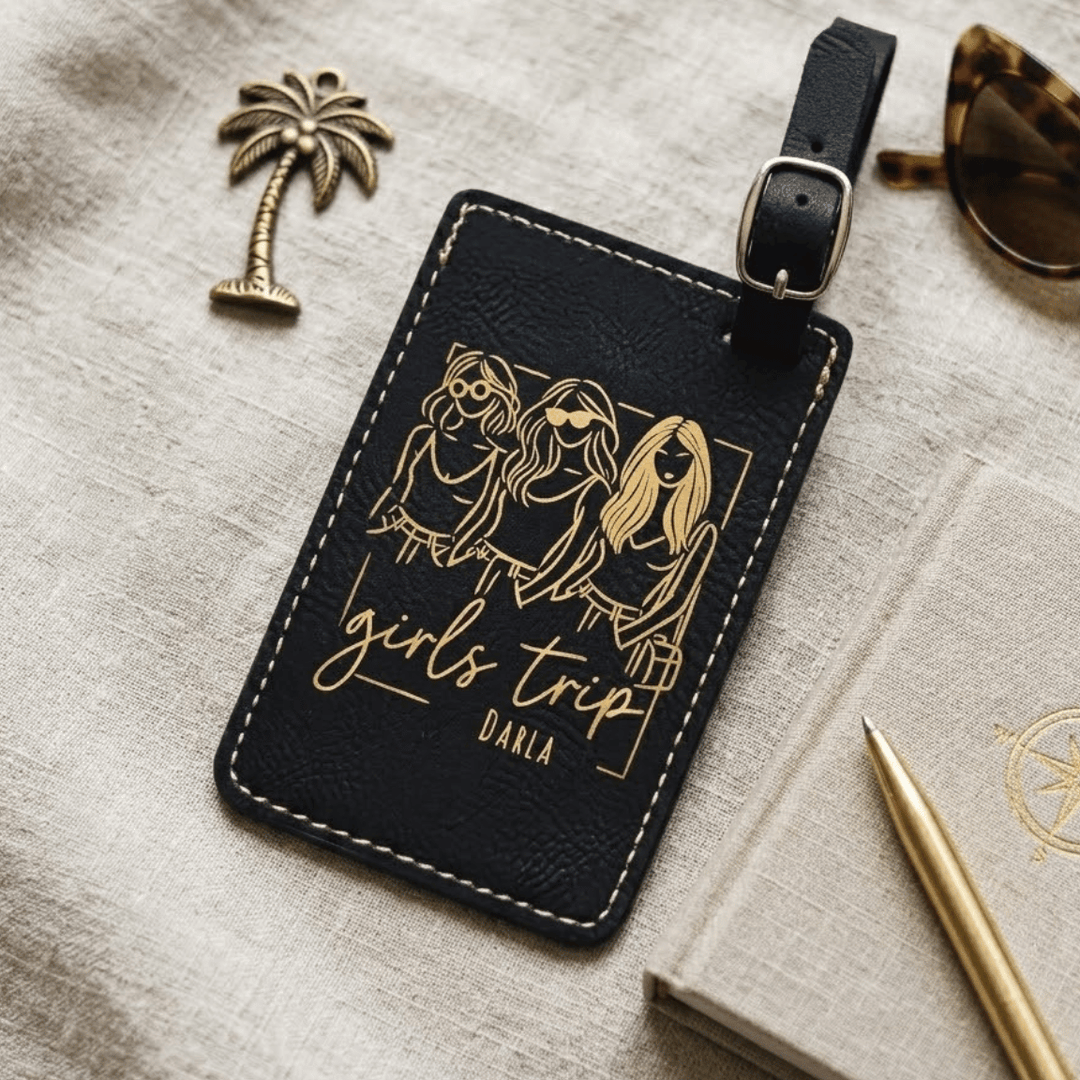 Black luggage tag with gold engraving of three women and 'girls trip' text, placed on a textured surface with accessories.