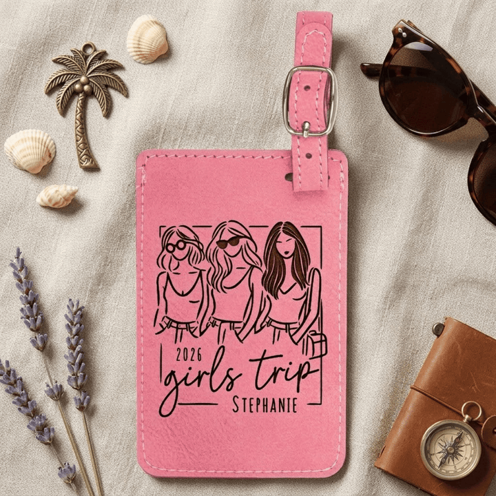 Pink luggage tag with '2026 Girls Trip Stephanie' design on a textured surface with travel-themed items.