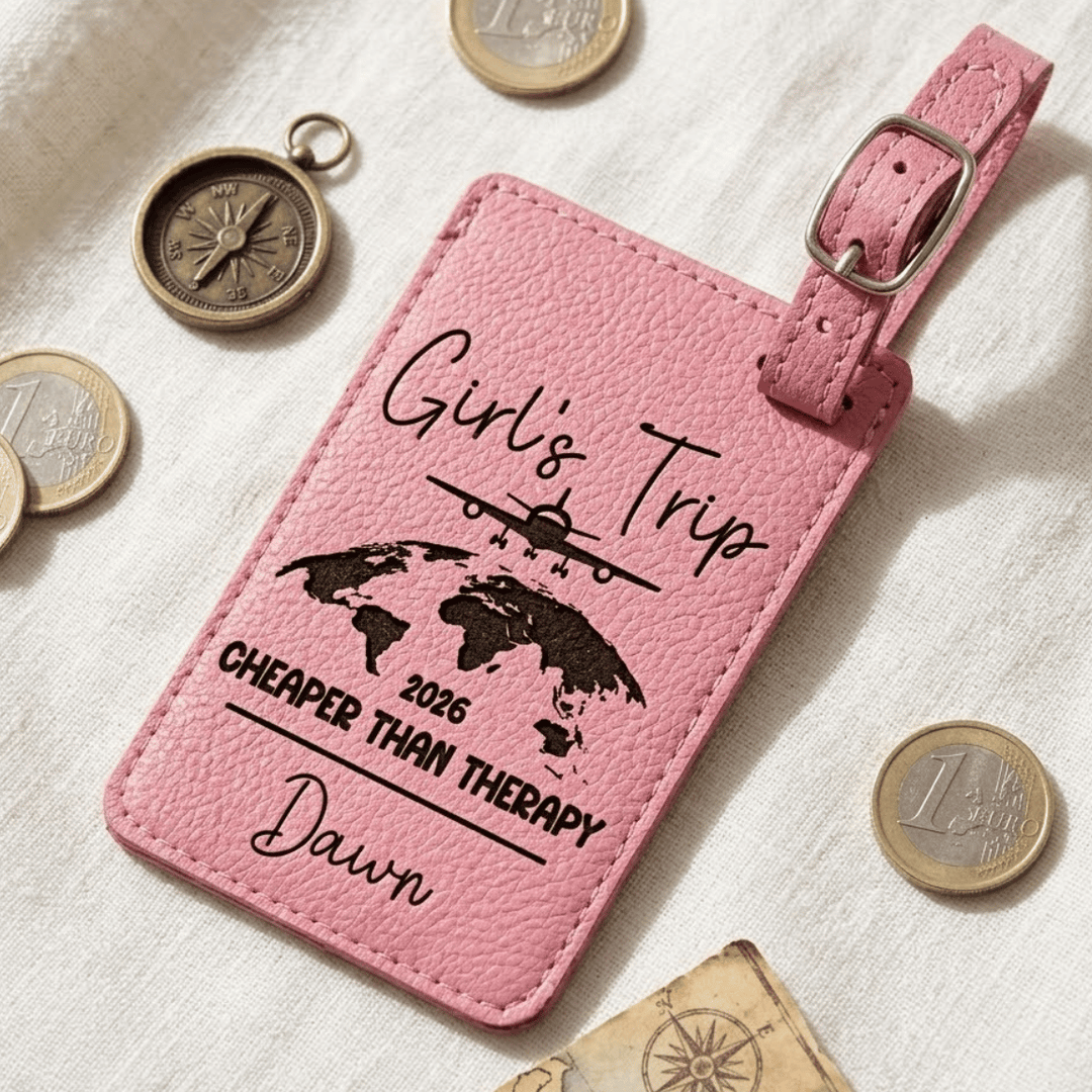 Pink luggage tag with 'Girl's Trip' text and world map design, surrounded by coins and a compass on a textured surface.