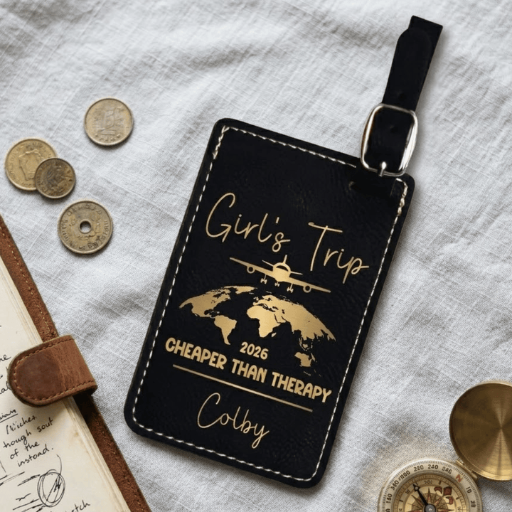 Black luggage tag with gold text and world map design on a textured surface with coins and a book.