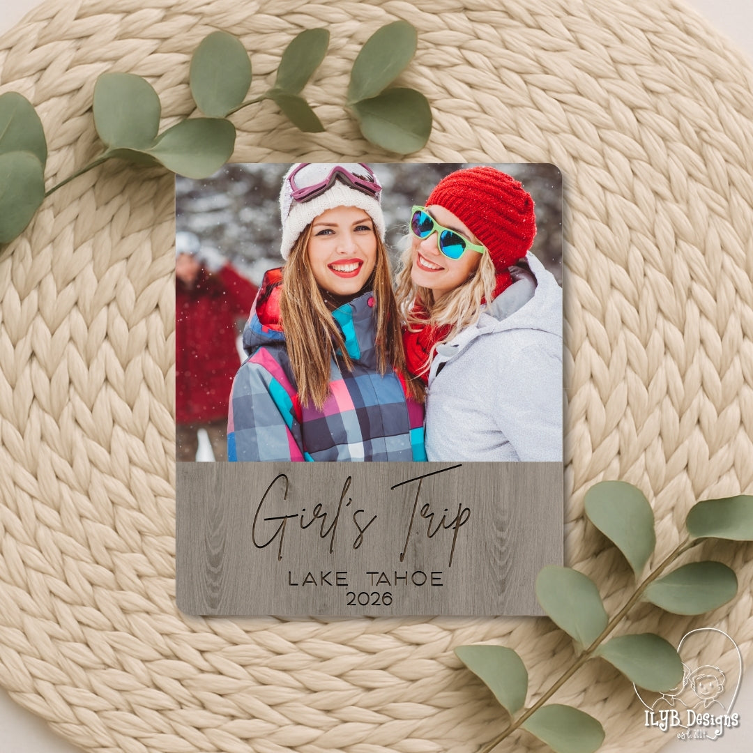 Travel photo board with a photo of two people and 'Girl's Trip Lake Tahoe 2026' text, on a textured surface with green leaves.