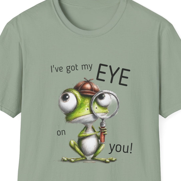 A sage green short-sleeve crewneck t-shirt featuring a cartoon graphic of a green frog wearing a magnifying glass and a brown hat, with the text 'I've got my EYE on you!' printed on the front.