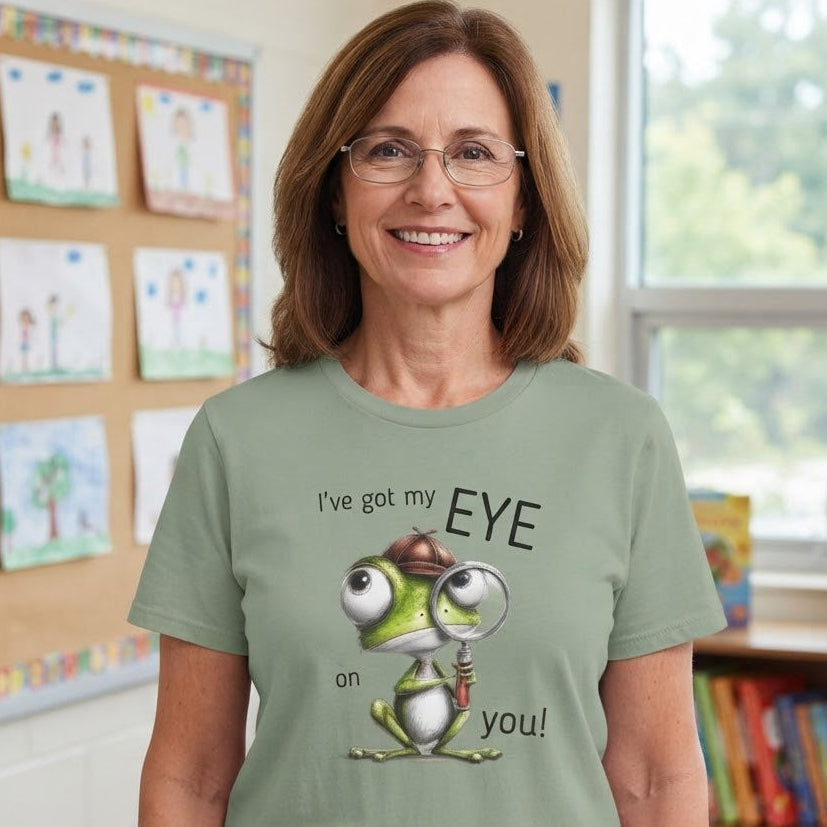 A woman wearing a sage green graphic t-shirt with a cartoon frog and the text 'I've got my EYE on you!' standing in a brightly lit classroom.