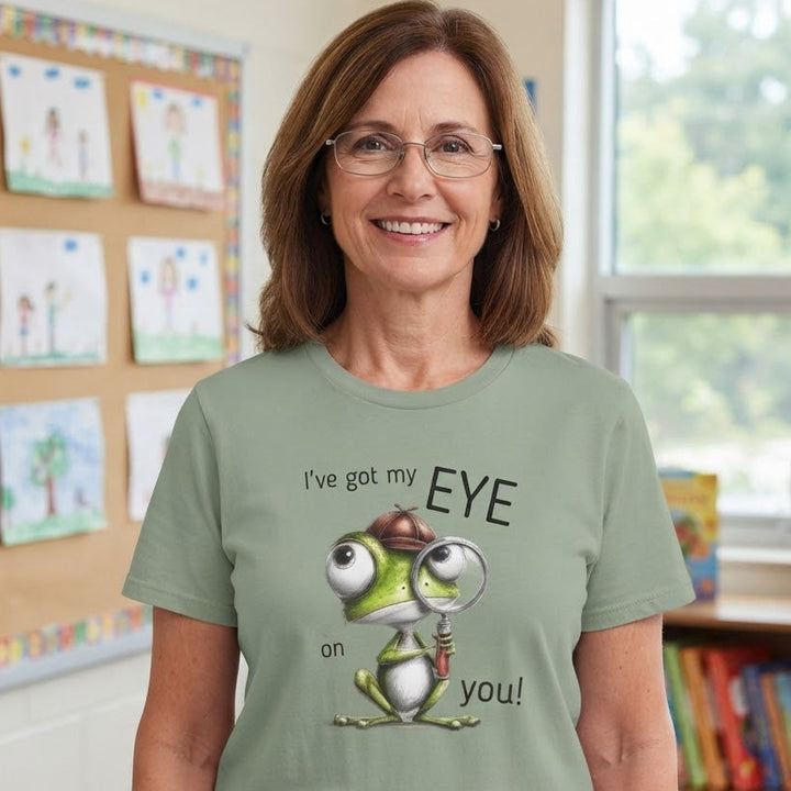A woman wearing a sage green graphic t-shirt with a cartoon frog and the text 'I've got my EYE on you!' standing in a brightly lit classroom.