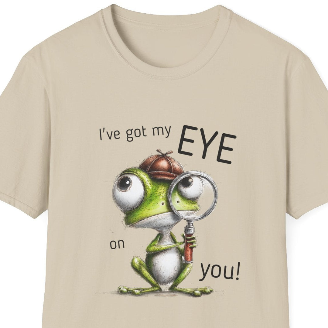 A beige short-sleeve crewneck t-shirt featuring a cartoon graphic of a green frog wearing a brown hat and magnifying glass, with the text 'I've got my EYE on you!' printed around it.