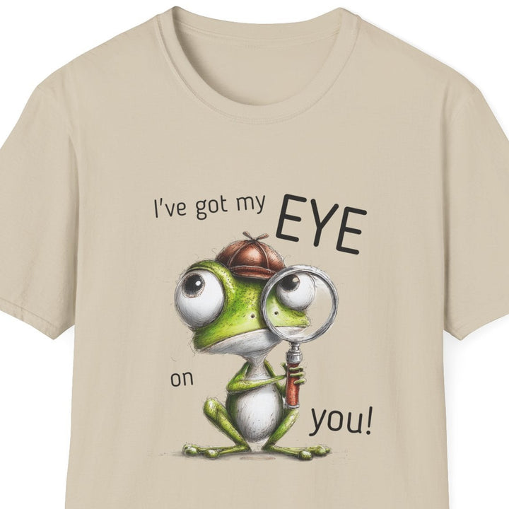 A beige short-sleeve crewneck t-shirt featuring a cartoon graphic of a green frog wearing a brown hat and magnifying glass, with the text 'I've got my EYE on you!' printed around it.