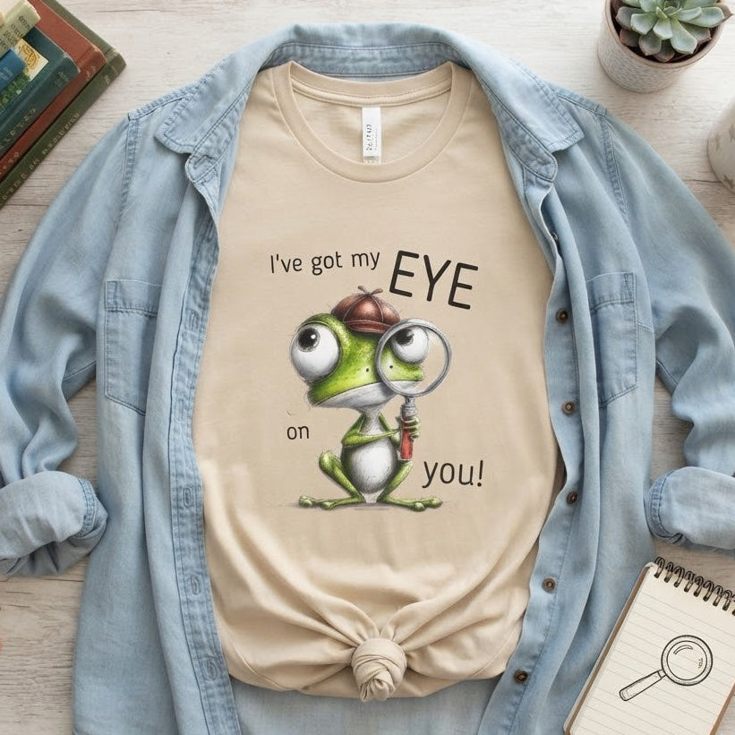 A beige graphic T-shirt featuring a cartoon frog with a magnifying glass and the text 'I've got my EYE on you!', styled in a flat-lay with an open light blue denim shirt layered over it, surrounded by books, a mug, a notebook, and a small succulent on a light wood surface.
