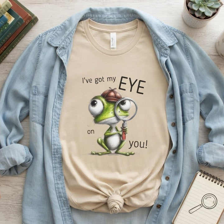 A beige graphic T-shirt featuring a cartoon frog with a magnifying glass and the text 'I've got my EYE on you!', styled in a flat-lay with an open light blue denim shirt layered over it, surrounded by books, a mug, a notebook, and a small succulent on a light wood surface.