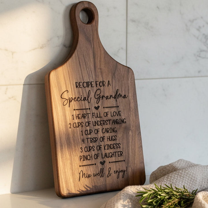 A personalized wooden cutting board with a handle, featuring an engraved recipe for a special grandma leaning on a white kitchen backsplash.