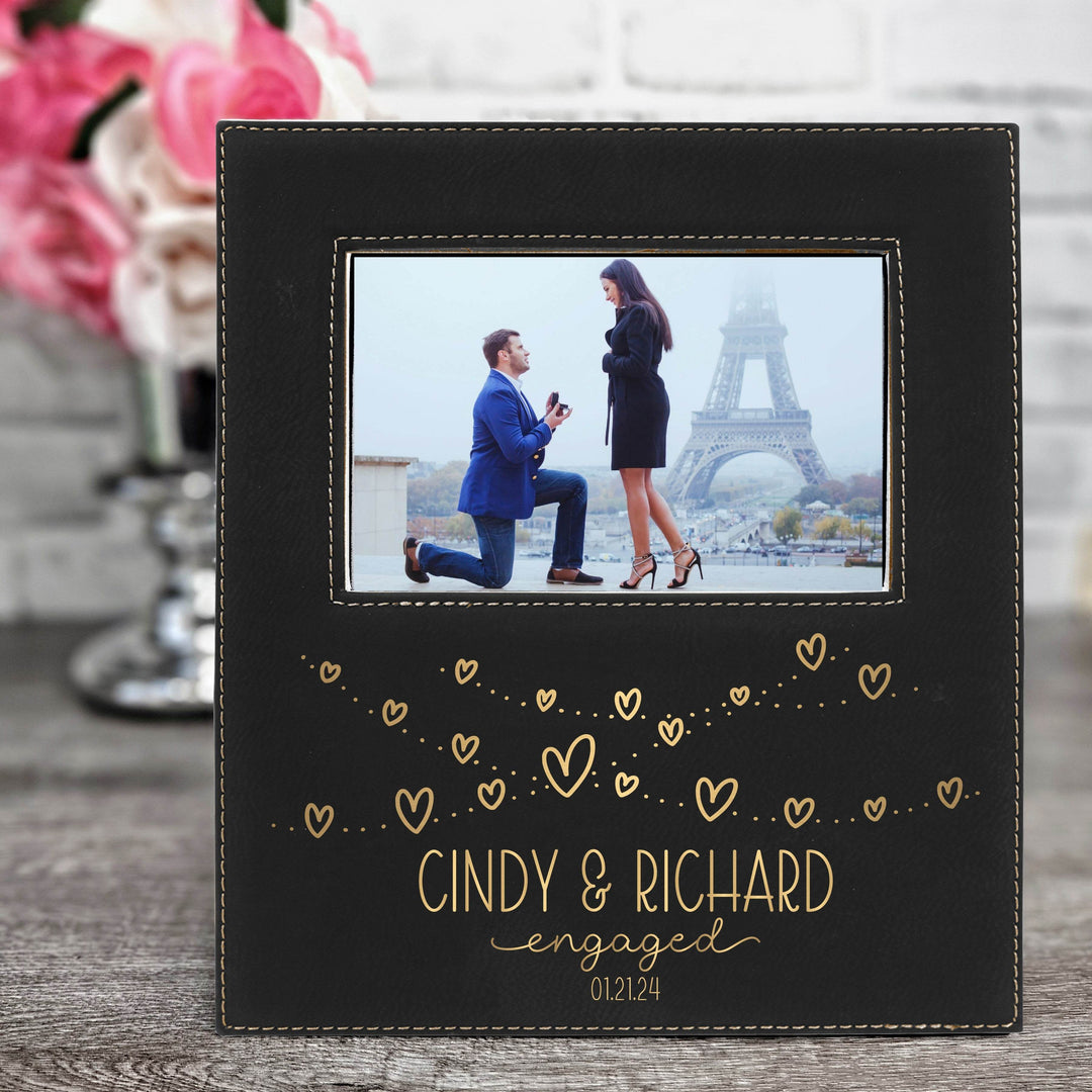 Personalized black vegan leather engagement photo frame with engraved names and text 'Cindy & Richard engaged 01.21.24' in metallic gold on a wooden surface.